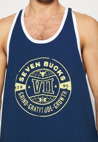 Granatowy podkoszulek z białym wykończeniem, z okrągłym emblematem w kolorze żółtym. Na podkoszulku znajduje się napis "SEVEN BUCKS", "VII" oraz "GRIND. GRATITUDE. GROWTH."