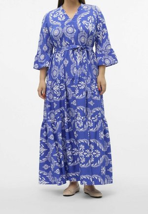 Vrouw draagt een lange blauwe jurk met bloemenpatroon, 3/4 mouwen met ruches, een ceintuur in de taille, en beige ballerina's.