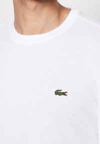 Vit bomullst-shirt med rund halsringning, med en liten grön broderad krokodillogo på vänster bröst.