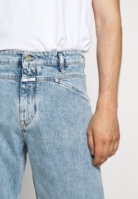 Jeans de mezclilla azul claro con cintura alta, con un cierre de botón, diseño clásico de cinco bolsillos y un desgastado visible para dar textura.