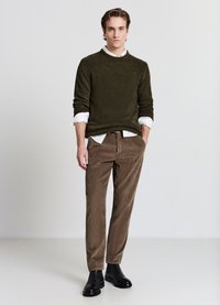 Olivgrüner strukturierter Pullover über einem weißen Hemd, kombiniert mit braunen Cordhosen und schwarzen Stiefeletten, vor einem schlichten Hintergrund.