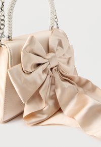 Μπεζ σατέν τσάντα χειρός με υπερμεγέθη φιόγκο και λαβές διακοσμημένες με μαργαριτάρια, ακουμπισμένη σε απλή λευκή επιφάνεια.