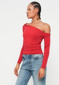 Czerwony top na ramiączkach z długimi rękawami, zmarszczoną strukturą i węzłem na ramieniu. Połączony z jasnoniebieskimi dżinsami.