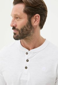 Weißes Langarmshirt mit Rundkragen, drei dunklen Knöpfen und einem kleinen gestickten Logo auf der Brust. Weicher, strukturierter Stoff.