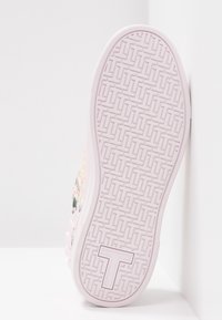 Semelle de sneaker rose clair avec un motif texturé, présentant un logo "T" bien visible et un design floral sur le côté. Matériau en caoutchouc lisse.