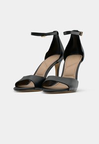 Black high-heeled sandals with ankle straps and open toes, beige cushioned insoles, displayed on a white background.