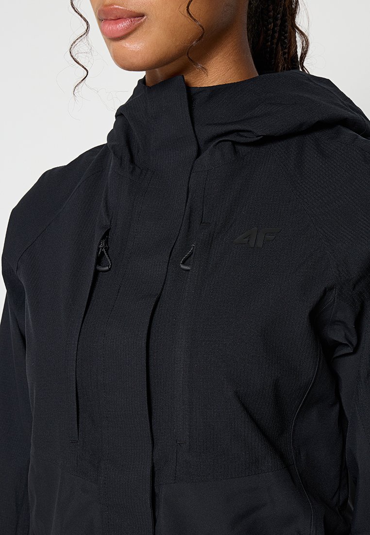 Zbliżenie na osobę w czarnej kurtce z kapturem, z zamkiem błyskawicznym i logo na piersi.