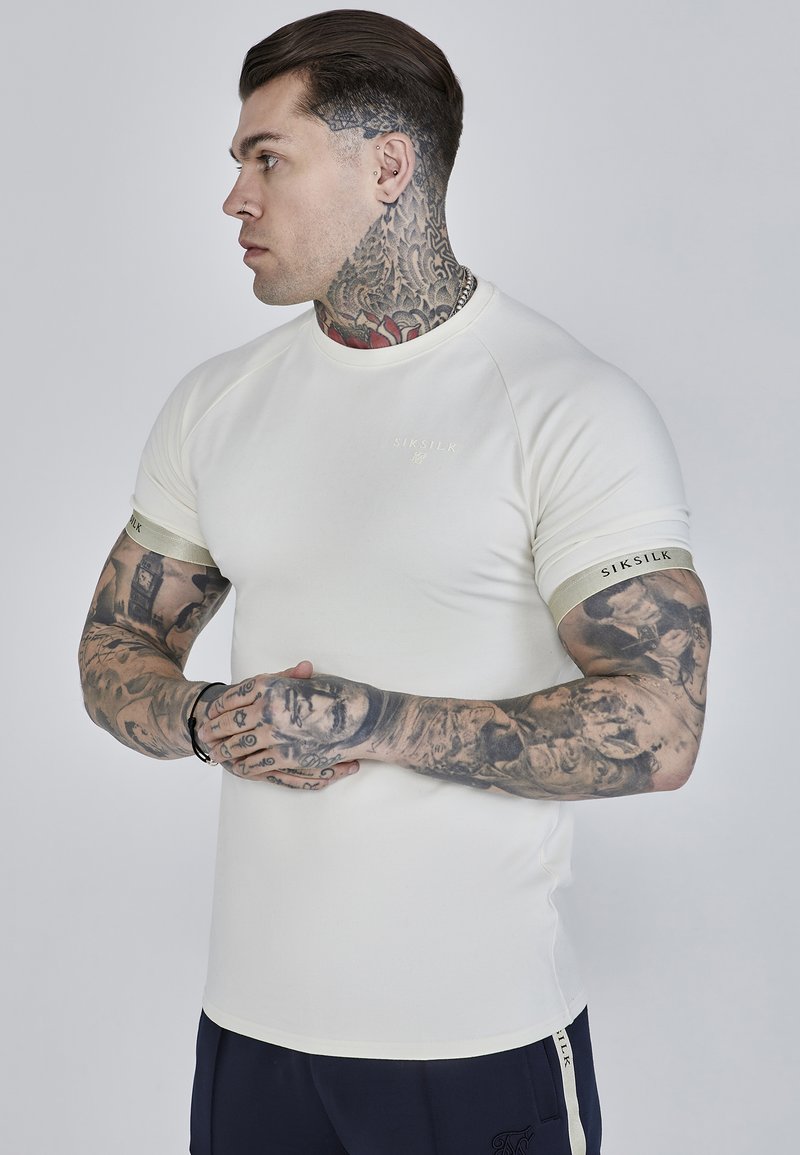 Beige lyhythelma T-paita, jossa on lyhyet raglan-hihat, kultainen logo rinnassa ja erottuva brändäys hihansuissa. Sileä pinta.