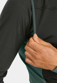 Langarm-Sportoberteil mit einem dunkelgrauen Körper und einem grünen Seitenpanel sowie strukturiertem Stoff. Verstellbarer Riemen auf der Schulter sichtbar.