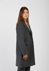 Donna con lunghi capelli castani che indossa un cappotto grigio scuro fino al ginocchio e posa di lato su uno sfondo chiaro e semplice.