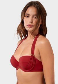 Červený bikini top s kosticemi, s nařasenou tkaninou a zlatými kovovými akcenty. Obsahuje široké popruhy a strukturovaný design. Hladká textura.