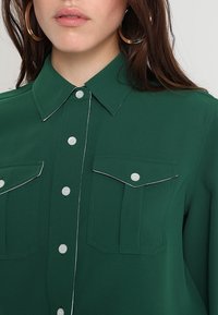 Camicia verde con colletto, due tasche sul petto e cuciture bianche a contrasto. Il tessuto ha una texture liscia.