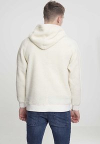 Man som bär en krämfärgad fluffig huvtröja och blå jeans, vänd bort från kameran mot en enkel ljus bakgrund.