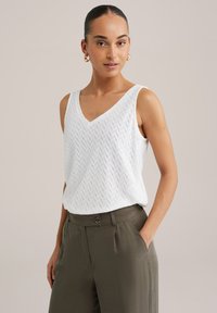 Bílý bezramenný top s výstřihem ve tvaru V, se strukturovaným vzorem herringbone, doplněný olivově zelenými kalhotami s vysokým pasem.