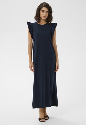 Žena s kučeravými vlasmi oblečená v tmavomodrých maxi šatách bez rukávov s volánikmi na ramenách a čiernymi sandálmi s otvorenou špičkou, stojaca pred jednoduchým pozadím.