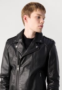 Young person with short light brown hair wearing a black leather jacket, looking sideways against a plain light background.