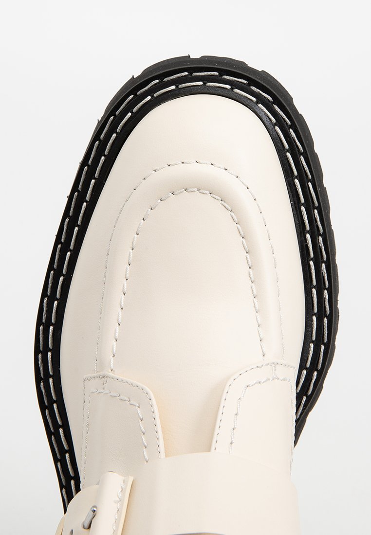Skórzane loafery w kolorze kremowym z wyraźną czarną gumową podeszwą, białymi detalami przeszycia i gładką fakturą. Charakteryzują się smukłym, zaokrąglonym kształtem.