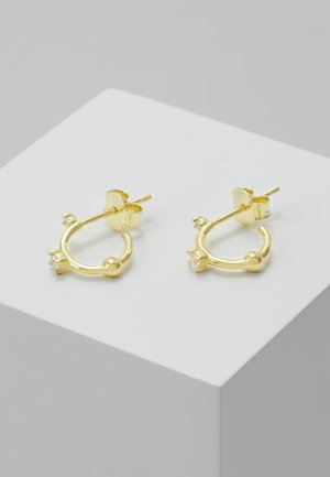 Petites boucles d'oreilles créoles en or avec une seule pierre précieuse claire et un détail en forme de boule, présentées sur un socle blanc sur un fond gris.