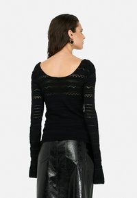 Black long-sleeve top featuring an open back, textured knit design with patterns, and fitted silhouette, paired with a shiny black skirt.