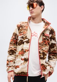 Ζακέτα φλις σε καφέ, κρεμ και πορτοκαλί αφηρημένο μοτίβο· διαθέτει κόκκινο φερμουάρ και λογότυπο Columbia. Το μοντέλο φοράει λευκό πουκάμισο από μέσα.