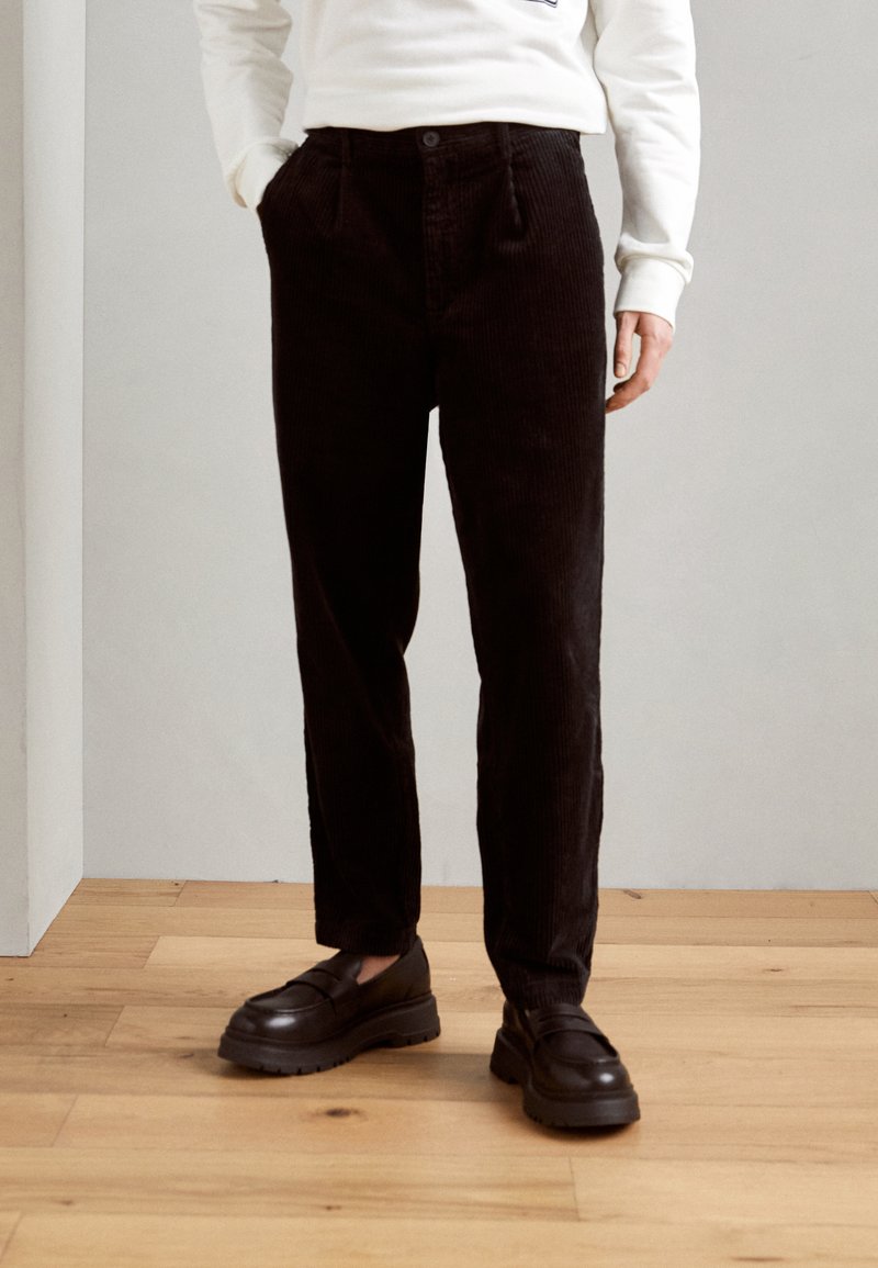 Pantaloni negri din catifea cu croi drept, dotati cu buzunare laterale si inchidere cu nasture, asortati cu loafers negri chunky pe o podea din lemn.