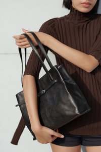 Oseba v rjavem rebrastem zgornjem delu oblačila, ki drži veliko črno usnjeno torbo z dvojnimi trakovi blizu telesa, nevtralen ozadje.