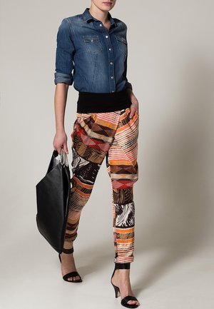 Vrouw met een denim blouse en kleurrijke broek met patroon, houdt een grote zwarte handtas vast en loopt op zwarte sandalen met hak.