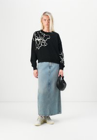 Felpa nera con motivo floreale bianco sulla spalla sinistra, abbinata a una gonna maxi in denim azzurro chiaro e scarpe da ginnastica bianche. Borsa nera in mano.