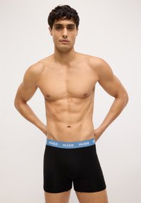 I boxer in cotone neri presentano un elastico azzurro chiaro con il logo "HUGO". Il modello ha un fisico tonico, con le braccia appoggiate ai fianchi.