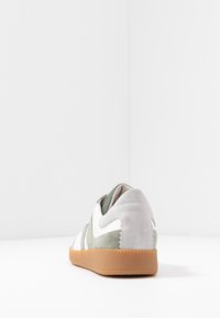 White and green suede sneaker with gum sole, viewed from the back on white surface against white background.