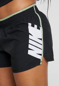 Zwarte sportshorts met een wit Nike-logo. Heeft een groene elastische tailleband en een zijsplit voor bewegingsvrijheid. Gemaakt van lichte materialen.