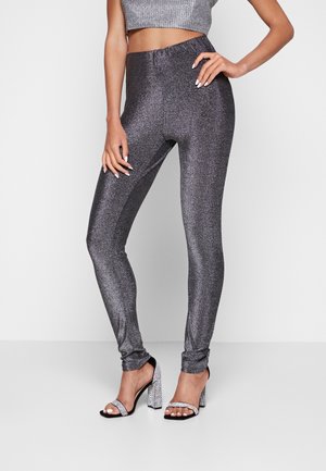 Glanzende, aansluitende, metallic grijze leggings met een elastische tailleband, gepaard met zilver belegde hoge hakken, tegen een neutrale achtergrond.