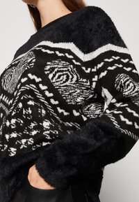 Femme portant un pull en peluche noir et blanc à motifs abstraits géométriques, les mains dans les poches d’un pantalon en cuir noir.