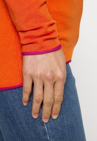 Maglione lavorato a maglia arancione con bordi viola ai polsini, indossato su denim blu. Mostra una mano appoggiata all'anca per contesto.