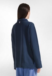 Granatowo-niebieski, oversize'owy sweter wykonany z miękkiego materiału, z kołnierzykiem i małą metką z logo z tyłu przy dekolcie. Prosty design, bez wzorów.