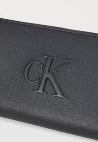 Portefeuille en cuir noir avec le logo "CK" estampé, présentant une texture lisse et un design épuré et minimaliste. Bords soigneusement cousus.