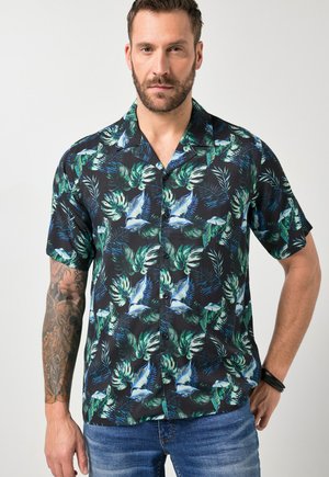 Mann mit Bart und Tätowierung, der ein schwarzes kurzärmeliges Hemd mit grünem Blätter- und weißem Vogelmuster sowie blaue Jeans trägt.