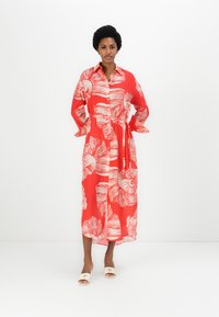 Rochie căștie cu model roșu și talie legată, având desene albii și nasturi în față. Pur tată cu sandale de tip slide în culori deschise.