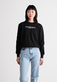 Sudadera negra con el logo blanco de "Calvin Klein Jeans", cuello redondo y mangas largas, combinada con unos pantalones vaqueros de tiro alto en un tono azul claro.