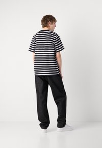 Tricou alb cu dungi negre asociat cu blugi închiși, cu croi lejer, și adidași albi. Materialul pare moale, având o croială relaxată.