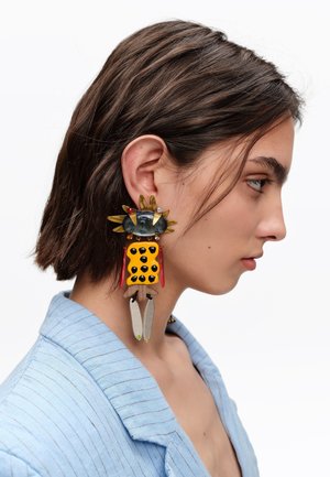 Joven con cabello corto castaño que lleva un gran pendiente colorido con forma de figura estilizada, vestida con un blazer azul claro.