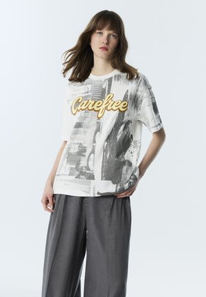 Młoda kobieta o brązowych włosach nosi luźny biały t-shirt z napisem „Carefree” i szerokie szare spodnie, stoi na jednolitym tle.