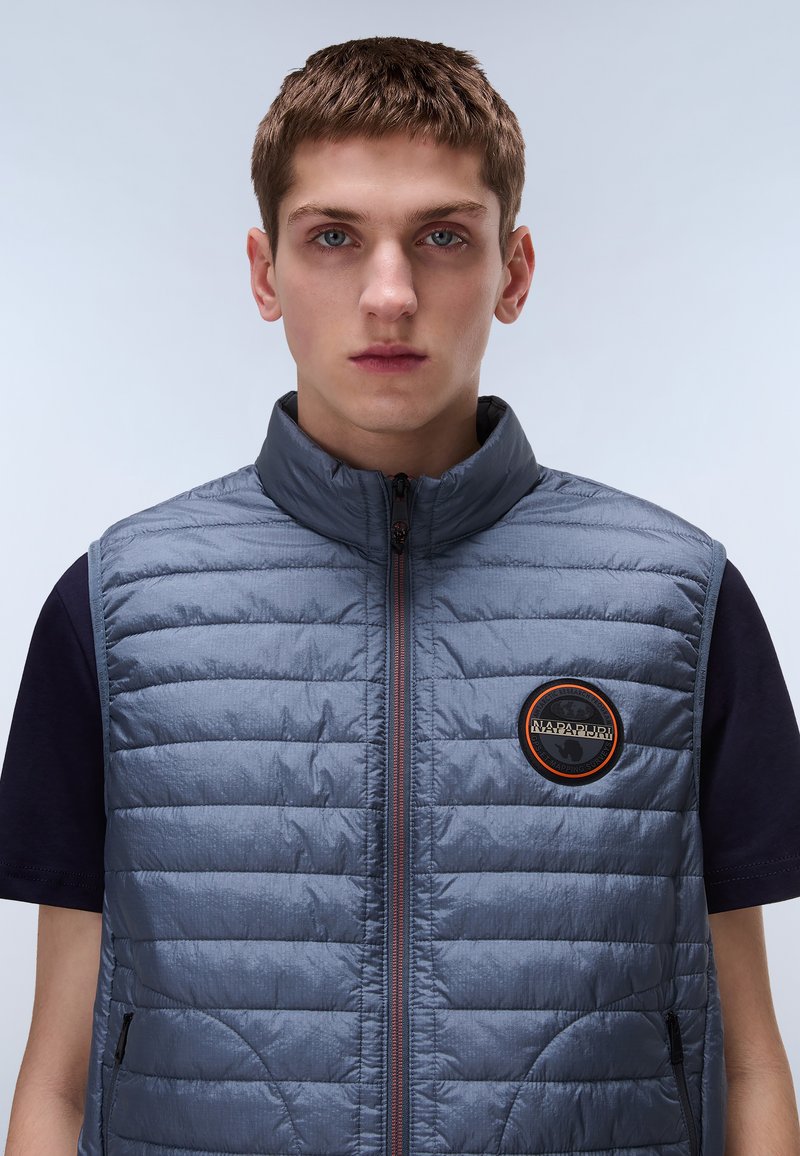 Párnázott szürke mellény magas gallérral, elülső cipzárral, narancssárga díszített logóval és oldalsó zsebekkel, sima, texturált felület jellemzi.