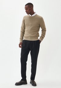 Suéter de punto color beige con cuello redondo, combinado con pantalones negros. El modelo lleva zapatos oscuros, exhibiendo una silueta relajada y ajustada.
