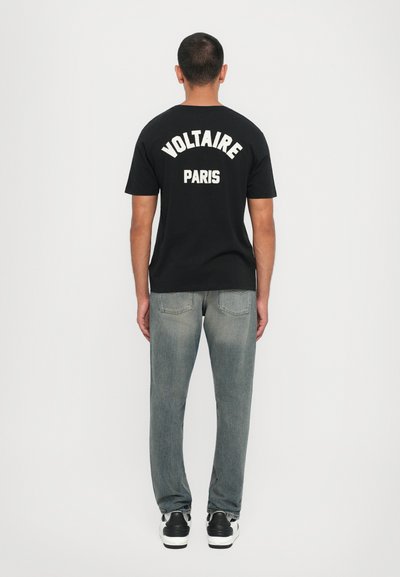 Svart t-skjorte med "VOLTAIRE" og "PARIS" i hvit tekst på baksiden, sammen med falmede grå jeans og svarte sneakers.