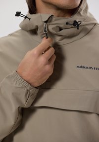 Veste beige imperméable avec un col élevé, fermeture éclair mi-hauteur, poignets élastiques et cordons de serrage réglables. Présente un logo sur la poitrine.
