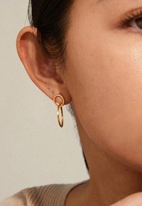 Detailní záběr na ucho osoby s krátkými tmavými vlasy, která má na sobě zlatý dvojitý kroužkový náušnici na neutrálním pozadí.