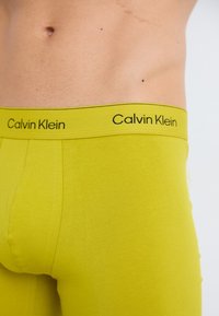 Calvin Klein Boxershorts aus leuchtend limettengrüner Baumwolle mit einem schwarzen Logo-Gummibund, die eine glatte Textur und eine enganliegende Form aufweisen.