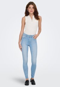 Uski bijeli rebrasti top bez rukava s patentnim zatvaračem, upareni sa svijetloplavim uskim trapericama i crnim slip-on cipelama.