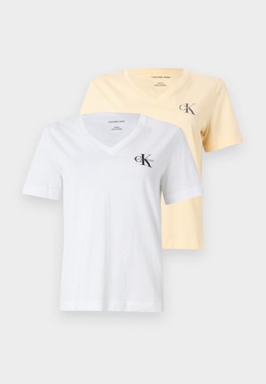 Kobieta stojąca przy białej ścianie, ubrana w żółtą koszulkę z dekoltem w serek, szerokie szare spodnie i jasnoróżowe baleriny, z rękami w kieszeniach.